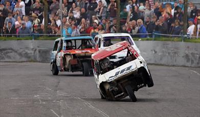 A three-wheeled banger racing car with one of its wheels in the air as it comes down the home straight