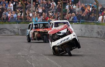A three-wheeled banger racing car with one of its wheels in the air as it comes down the home straight
