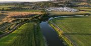 Overhead shot of a winding river cutting between two fields with hills and a caravan park behind