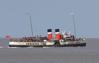 A paddle steamer out at sea with passengers crammed onto the deck
