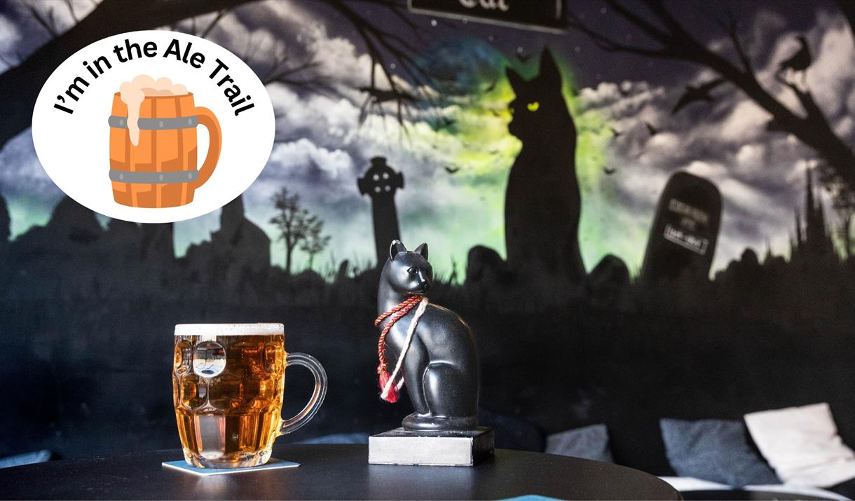 A pint of beer and a black cat set against a mural featuring a black cat and over-stamped with an I'm in the ale trail logo