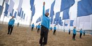 Actors dressed in blue t shirts and black trousers performing on a beach under blue and white flags