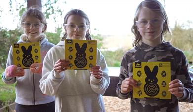 A group of children wearing protective glasses, holding up bright yellow Easter themed air rifle targets