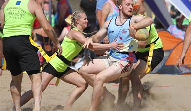 A woman in yellow attempts to tackle a man in blue during a Beach Rugby Festival