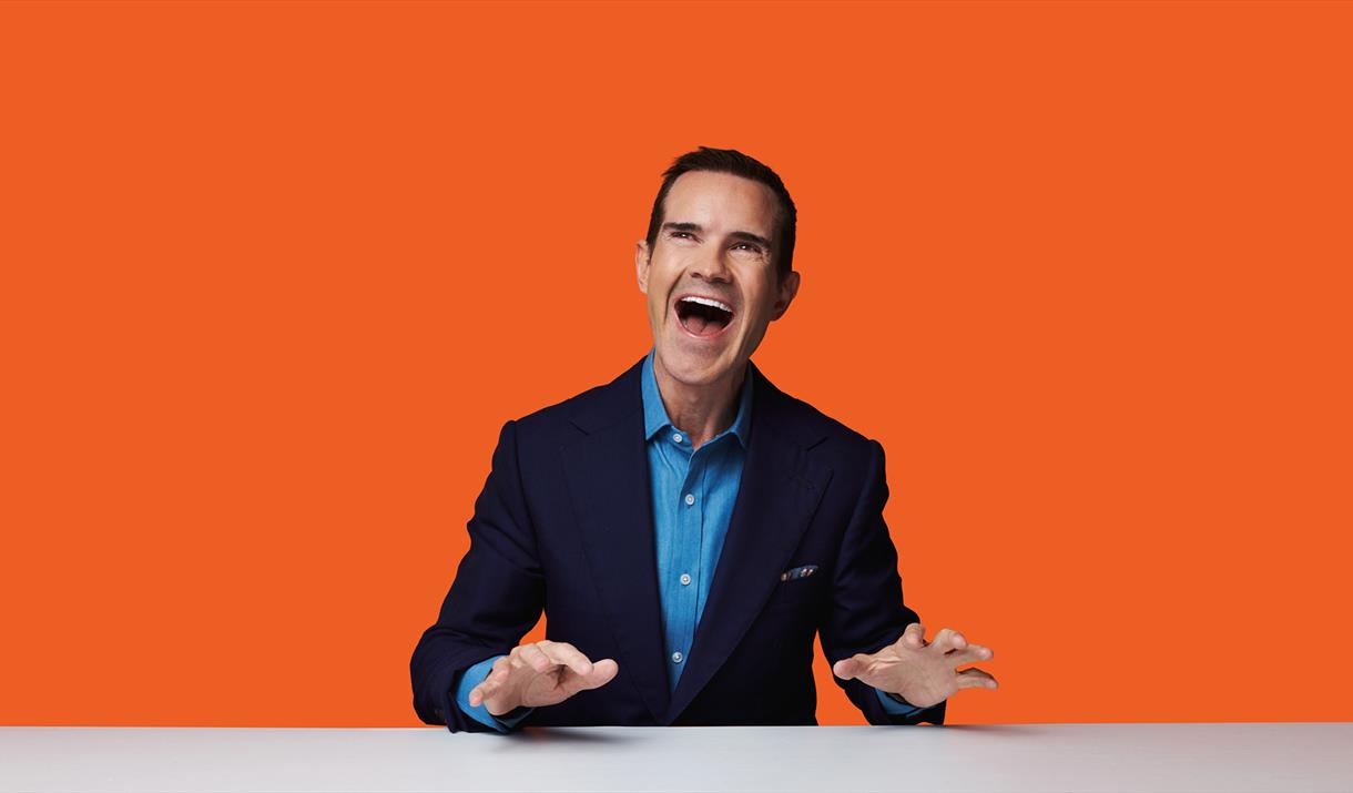 Photograph of Jimmy Carr stood against a orange background.