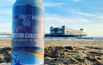 A  can of beer placed on a sandy beach with the sea and a pier in the background