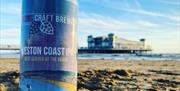 A  can of beer placed on a sandy beach with the sea and a pier in the background
