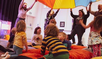 Bright photograph of children sitting on fluffy cushions under a bit coloured parachute.