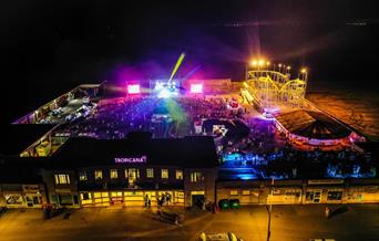 Aerial night time view of an entertainment venue with a funfair, bright stage lights and crowds of people