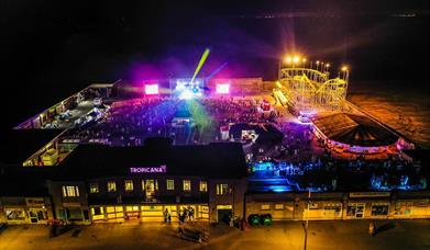 Aerial night time view of an entertainment venue with a funfair, bright stage lights and crowds of people