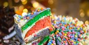 A slice of blue, green, red and white cake on top of the same cake covered in bright coloured Smarties sweets