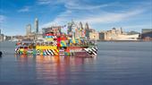 The Mersey Ferries brightly coloured Dazzle Ferry sailing on the Mersey River on a blue sky day. The iconic Liverpool skyline is behind the ferry.