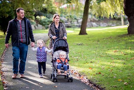 A man and woman hold hands with a young child. The Mother is also pushing a younger child in a pushchair. They are walking through a park.