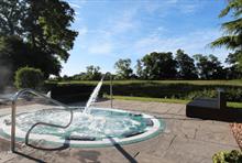 An outdoor sunken hot tub style pool with a large tap spraying water into the pool. Surrounding the pool is the countryside and lush grass.