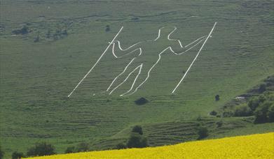 the outline of a white giant man carved in the side of the hill with two sticks surrounded by green countryside