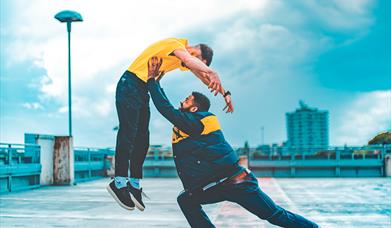 Two people dancing on the top level of a car park. One dancer is lifting the other up