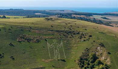 the outline of a white giant man carved in the side of the hill with two sticks surrounded by green countryside