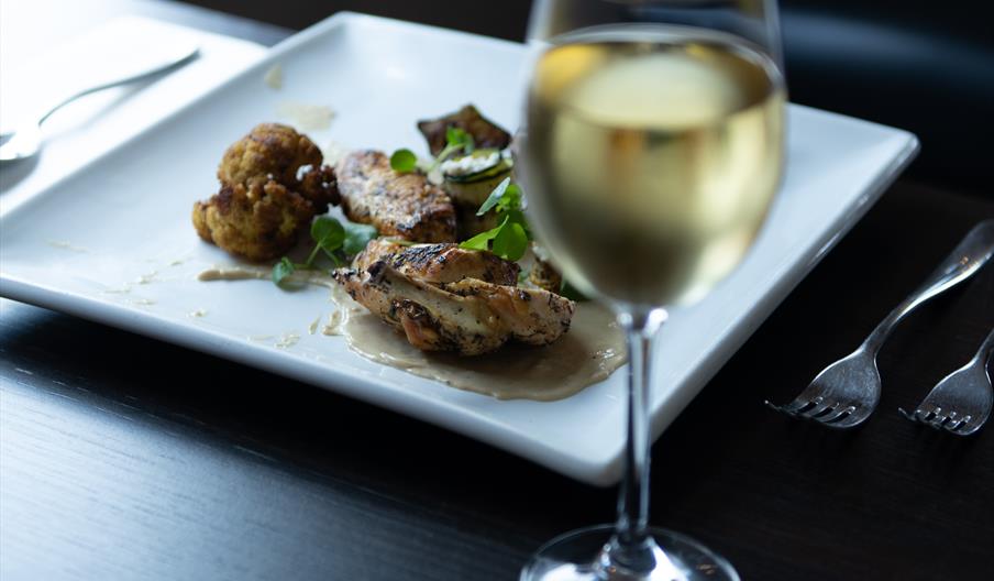 A photo of a wine glass in front of a plate of food on a square plate.