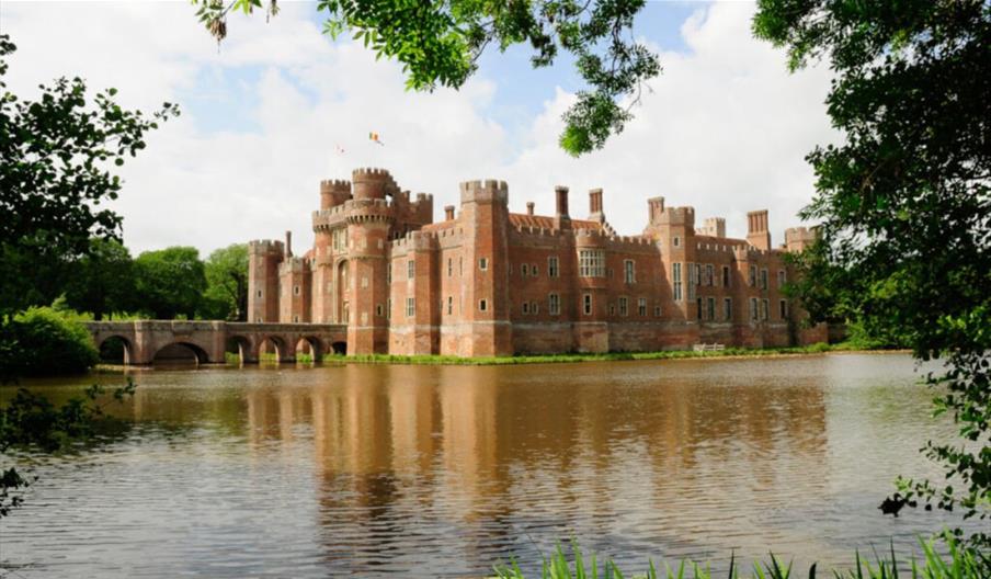 Red brick castle surrounded by a wide moat, with an arched stone bridge and reflections in the water, framed by green trees.