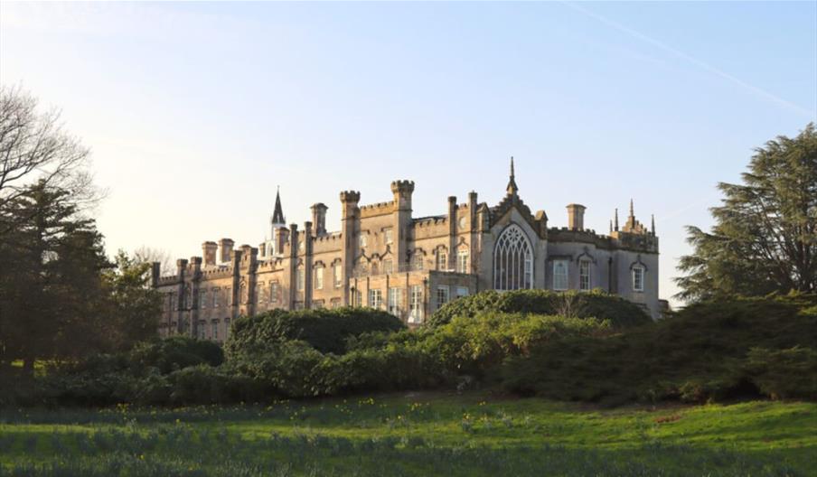 Historic country house with tall windows and decorative stonework, set behind landscaped gardens and trees in soft evening light.