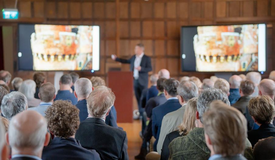 A person giving a lecture in a hall, with people watching