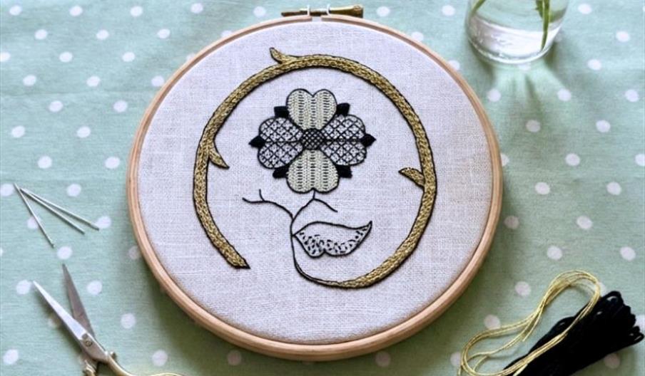 Embroidery hoop with an Elizabethan-style floral design in blackwork and gold braid stitching, surrounded by scissors, needles and thread on a pale gr