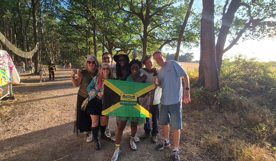Festivalgoers posing with a Jamaican flag along a woodland walkway at One Love Festival, surrounded by trees and colourful festival artwork.