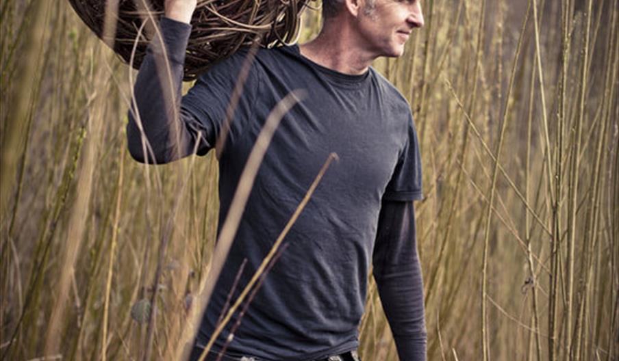 Basket Maker standing in a field with long reads holding a large woven ball of willow on his shoulder