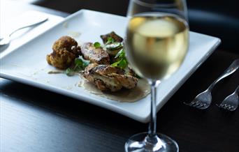 A photo of a wine glass in front of a plate of food on a square plate.