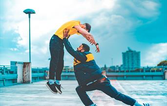 Two people dancing on the top level of a car park. One dancer is lifting the other up