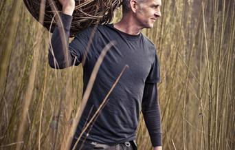Basket Maker standing in a field with long reads holding a large woven ball of willow on his shoulder