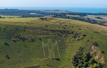the outline of a white giant man carved in the side of the hill with two sticks surrounded by green countryside