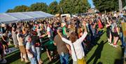 Large crowd gathered on a grassy field at an outdoor event, with people standing near a marquee tent and trees in the background.