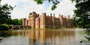Red brick castle surrounded by a wide moat, with an arched stone bridge and reflections in the water, framed by green trees.