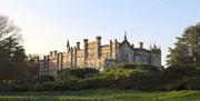 Historic country house with tall windows and decorative stonework, set behind landscaped gardens and trees in soft evening light.