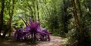 A large, purple, spiky creature puppet operated by performers stands on a woodland path surrounded by tall trees and dappled sunlight.