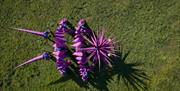 Aerial view of a large, purple, spiky puppet-like creature arranged across a grassy field, with long pointed limbs and a starburst-shaped head casting