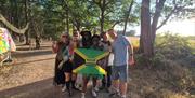 Festivalgoers posing with a Jamaican flag along a woodland walkway at One Love Festival, surrounded by trees and colourful festival artwork.