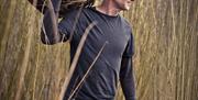 Basket Maker standing in a field with long reads holding a large woven ball of willow on his shoulder