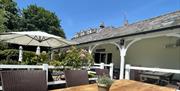 An outside terrace with a long wooden table and chairs around it. There is a large white parasol in the background with some flowers.