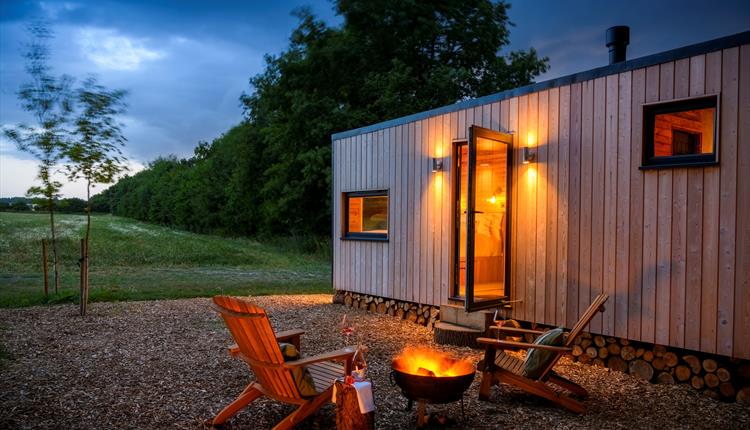 A photo of one of the cabins at dusk, showing the countryside view in the background, the glow of the firepit with 2 wooden armchairs around it and a