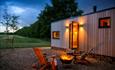 A photo of one of the cabins at dusk, showing the countryside view in the background, the glow of the firepit with 2 wooden armchairs around it and a