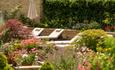 A line of cream sunbeds surrounded by Cotswold stone walls and pretty pink hues of flowers and plants.