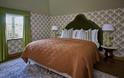 Cosy vintage bedroom with a rust-orange quilted bedspread, ornate wallpaper, green curtains, bedside lamps, and a patterned rug.