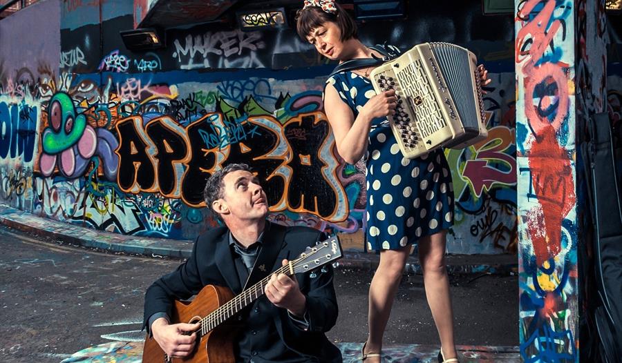 Oh la la! - Graham photo - woman playing accordion standing, man playing guitar sat, both in front of an urban graffitied landscape