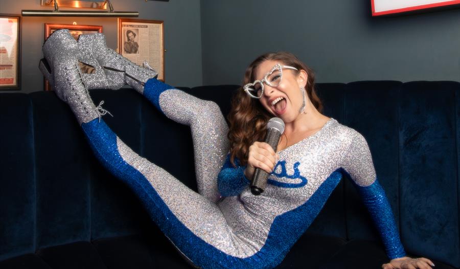 Jess Robinson: Elton Reimagined ©Chloe Hashem - women in sparkly outfit signing into a mic in a lounge with feet up