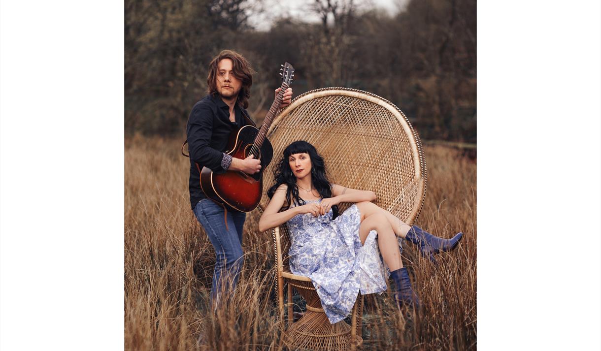 Hannah & Ben - Hannah sat in a wicker chair, Ben stood behind holding a guitar, in a field