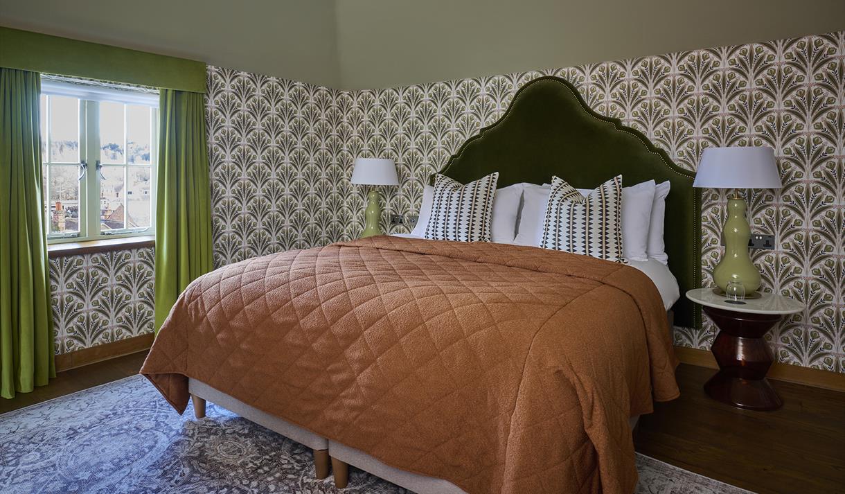 Cosy vintage bedroom with a rust-orange quilted bedspread, ornate wallpaper, green curtains, bedside lamps, and a patterned rug.