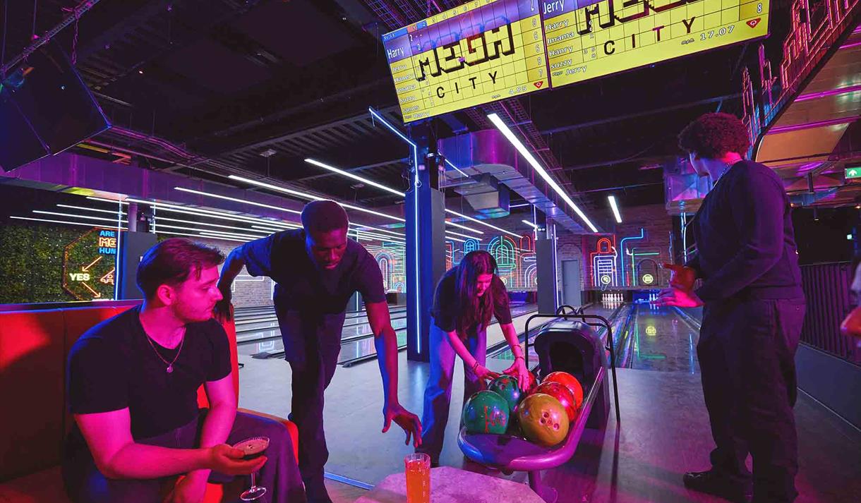 Four friends gather in a vibrant neon-lit arcade, examining colorful bowling balls in a cart beneath glowing “Mega City” signs.
