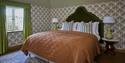 Cosy vintage bedroom with a rust-orange quilted bedspread, ornate wallpaper, green curtains, bedside lamps, and a patterned rug.